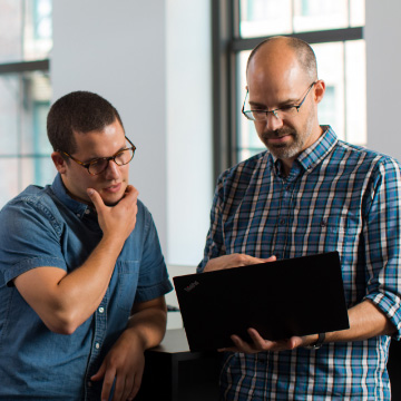 ISV-Entwickler arbeiten gemeinsam am Deployment einer App, beide Männer stehen in der Mitte eines Büros und schauen auf einen Computer